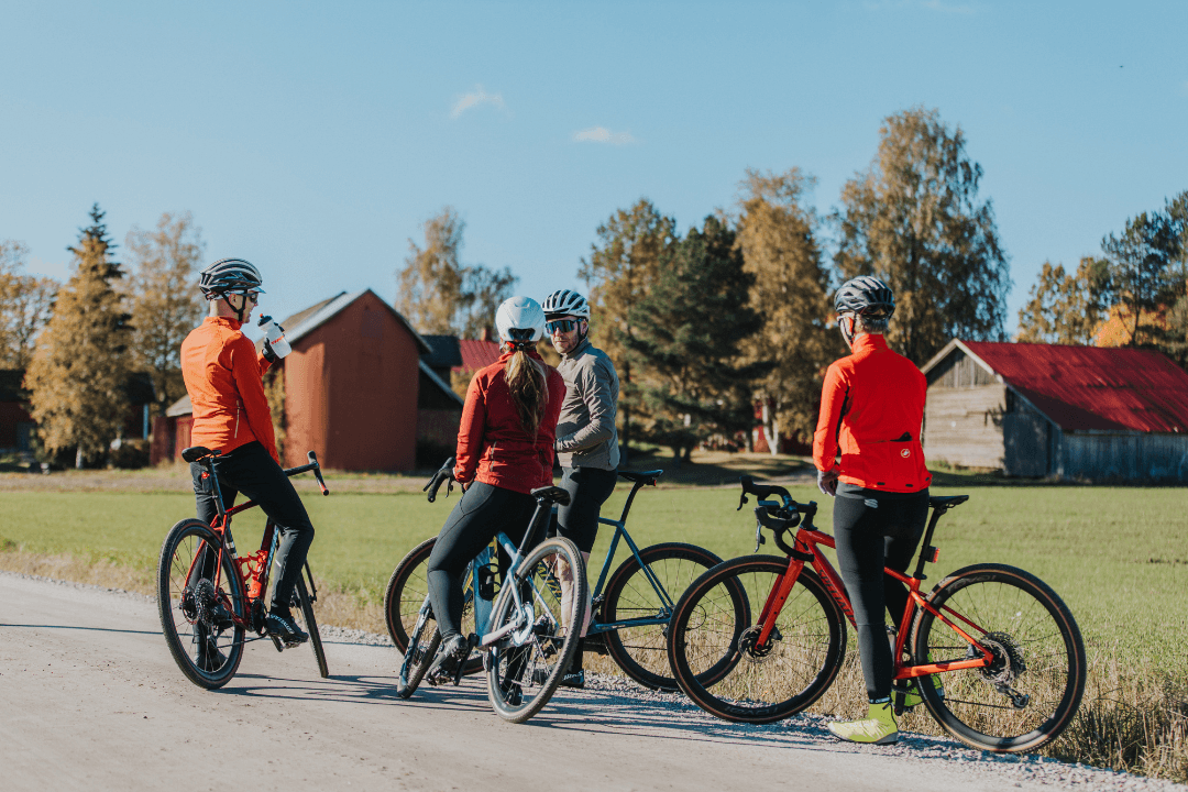 Four cyclists in kit paused on a rural gravel road with autumn trees and buildings in the background.